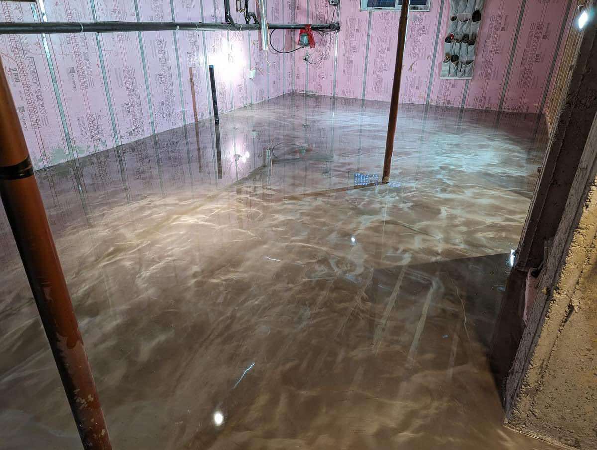 Sol époxy brillant et métallique dans un sous-sol. Isolation rose sur les murs. Deux poteaux et des tuyaux traversent l'espace.