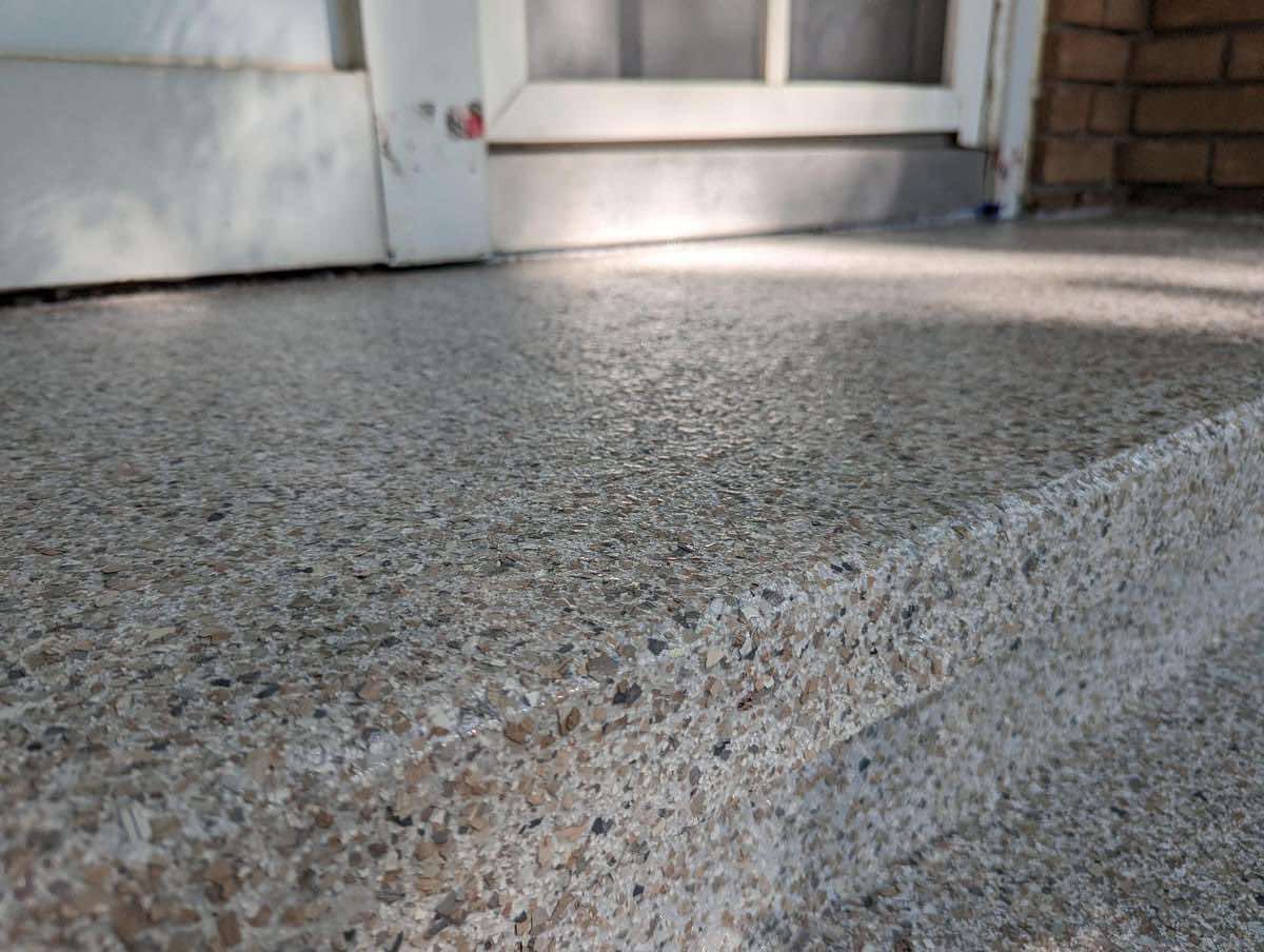 Gros plan d'une marche en béton gris avec une surface rugueuse et texturée devant une porte blanche.