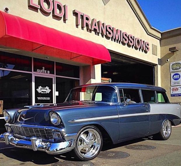 A classic blue and silver station wagon parked in front of Lodi Transmissions shop with a red awning.
