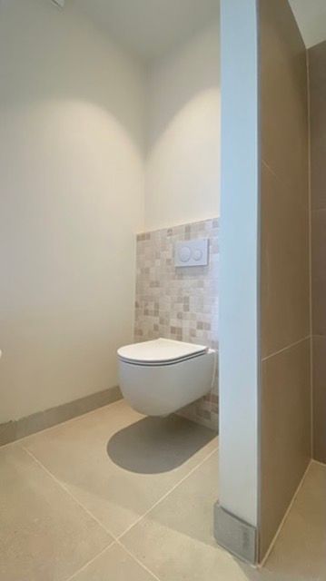 Een wit, aan de muur gemonteerd toilet in de badkamer. Lichtbruine tegelvloer en een neutrale kleur op de muren.