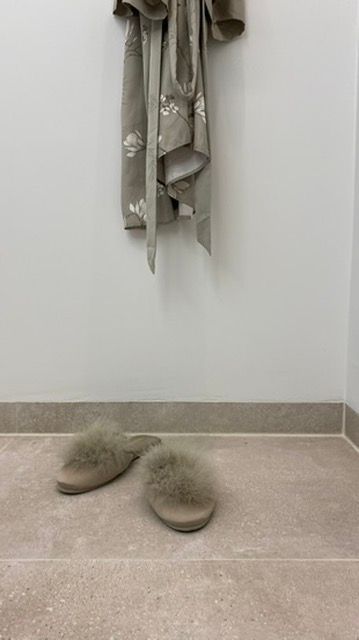 Een badjas hangt boven een paar pluizige slippers op een beige tegelvloer. Neutrale kleuren.