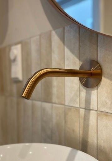 Wandkraan rose copper  kraan boven een witte wastafel, tegen een beige tegelwand en een ronde spiegel.