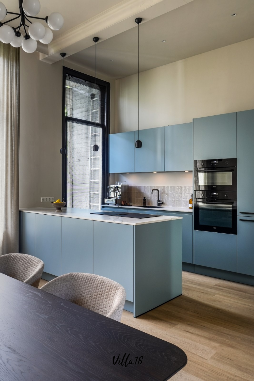 Lichtblauwe moderne keuken met kookeiland, zwarte apparatuur en hoog raam met houten woodblinds