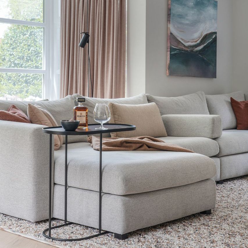 Gezellige woonkamer met een lichte hoekbank, een klein zwart tafeltje en een schilderij aan de muur.