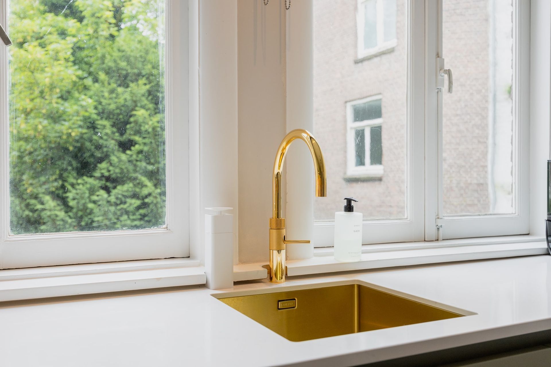 Gouden wasbak en kraan voor een raam met uitzicht op groen en een gebouw.