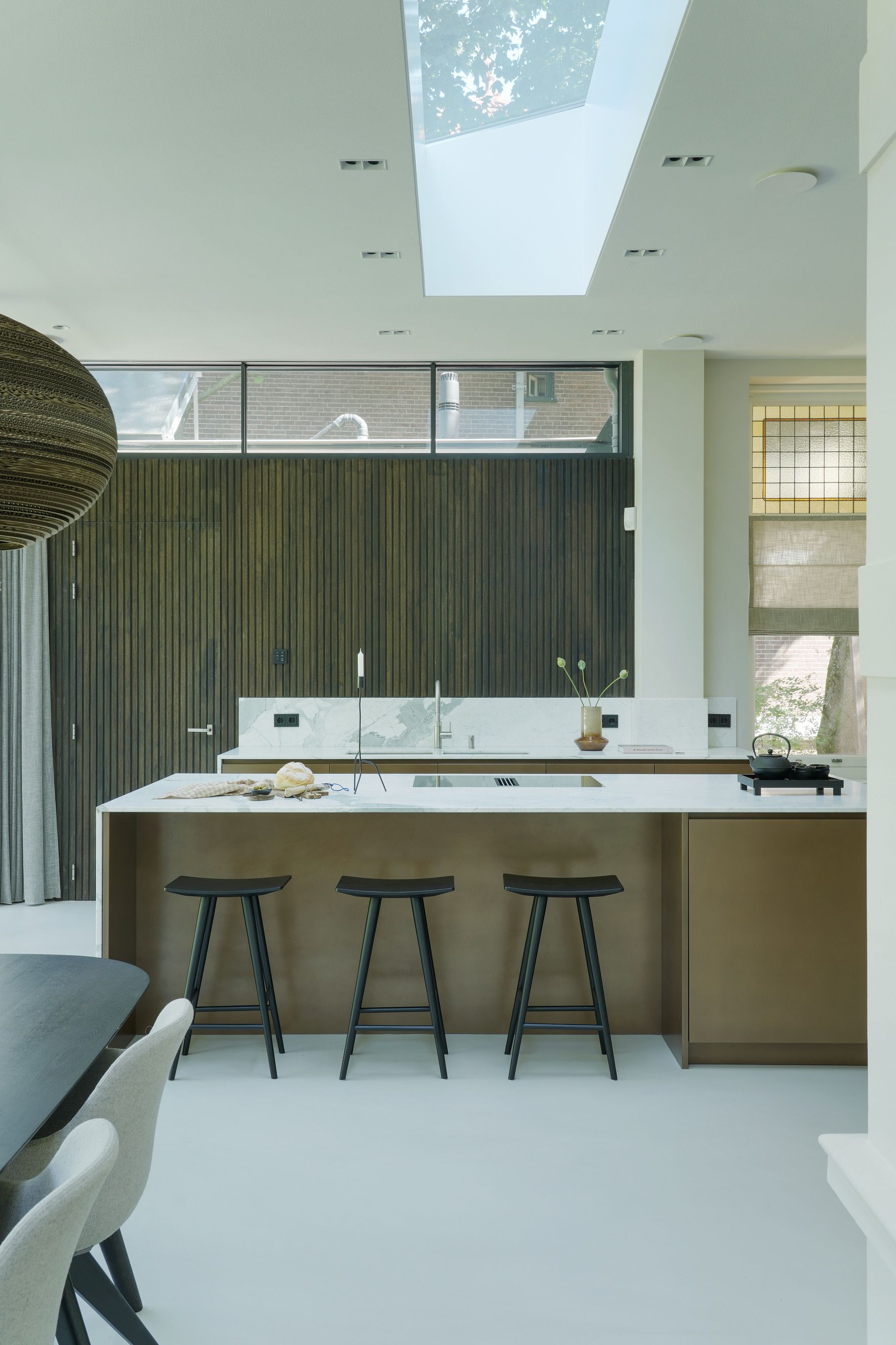 Moderne keuken met kookeiland, drie zwarte krukken en dakraam. Beige en bruine tinten domineren.