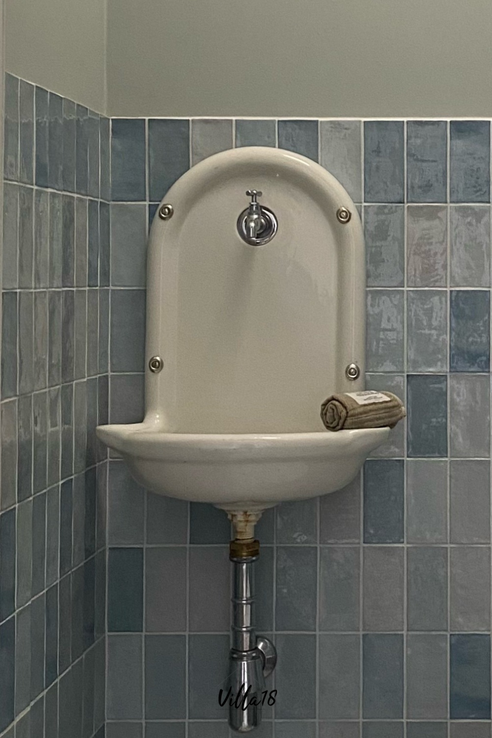 Witte, aan de muur gemonteerd antiek fonteintje met kraan en zeepbakje, tegen een muur van blauwe design tegels.