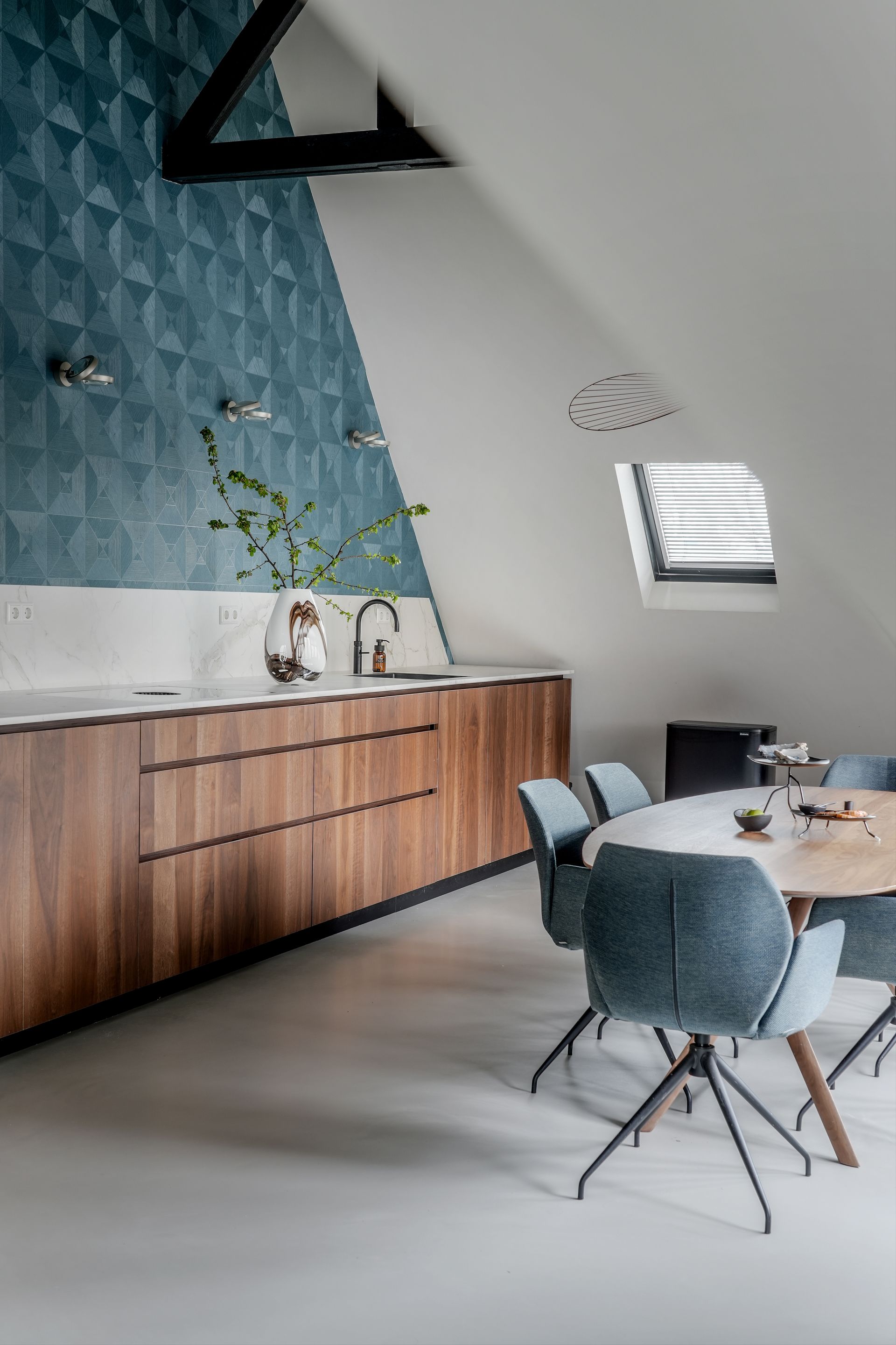 Keuken en eetkamer met geometrisch behang in blauwgroen, houten kasten en een ronde tafel met blauwe stoelen.