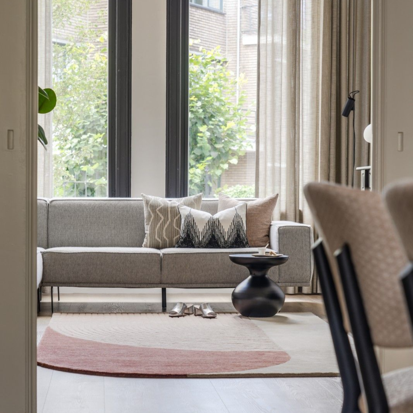 Moderne woonkamer met grijze bank, tapijt met patroon, grote ramen en klein zwart bijzettafeltje.