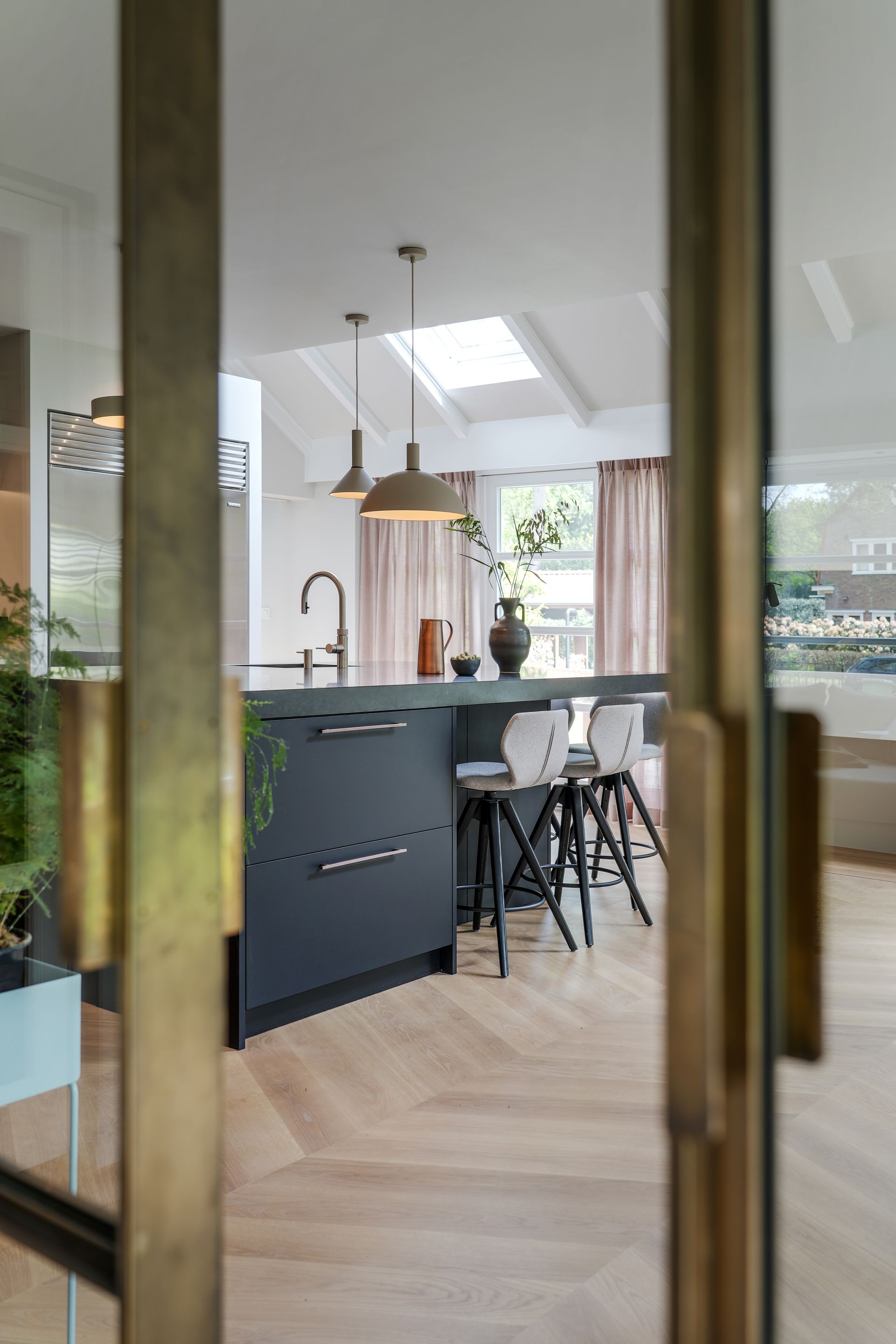 Keukeneiland met zwarte kasten, grijze krukken en lichte houten vloer, zichtbaar door glazen bronzen deuren.