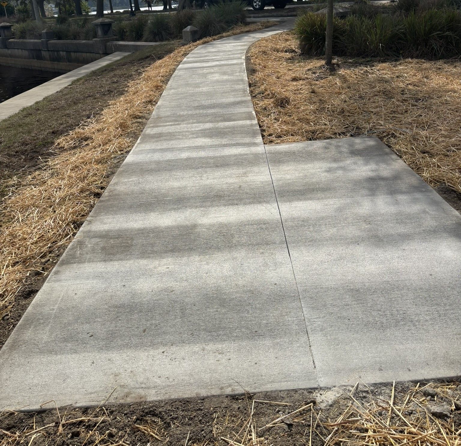 Concrete walkway curves along water's edge, past a bench and dry grasses.