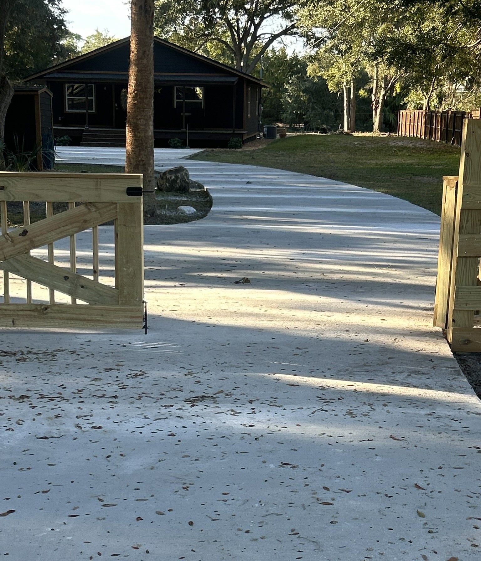 Wooden gate opens onto a concrete driveway leading to a dark house in a sunny outdoor setting.