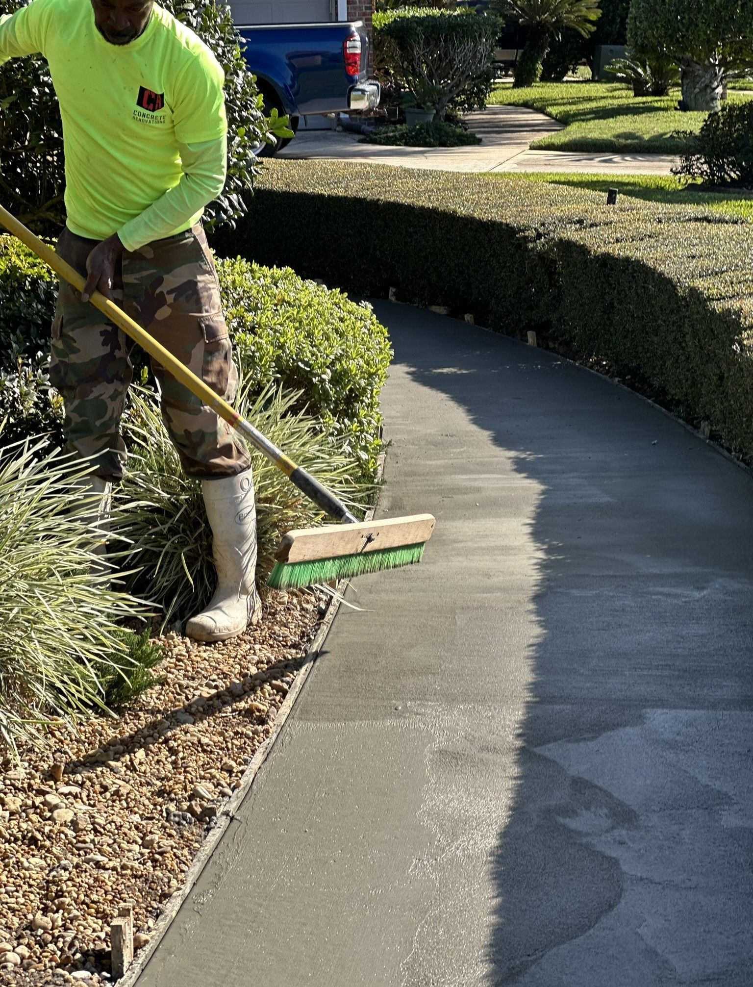 Person in camouflage pants and neon Concrete Renovations shirt brushing a wet sidewalk.