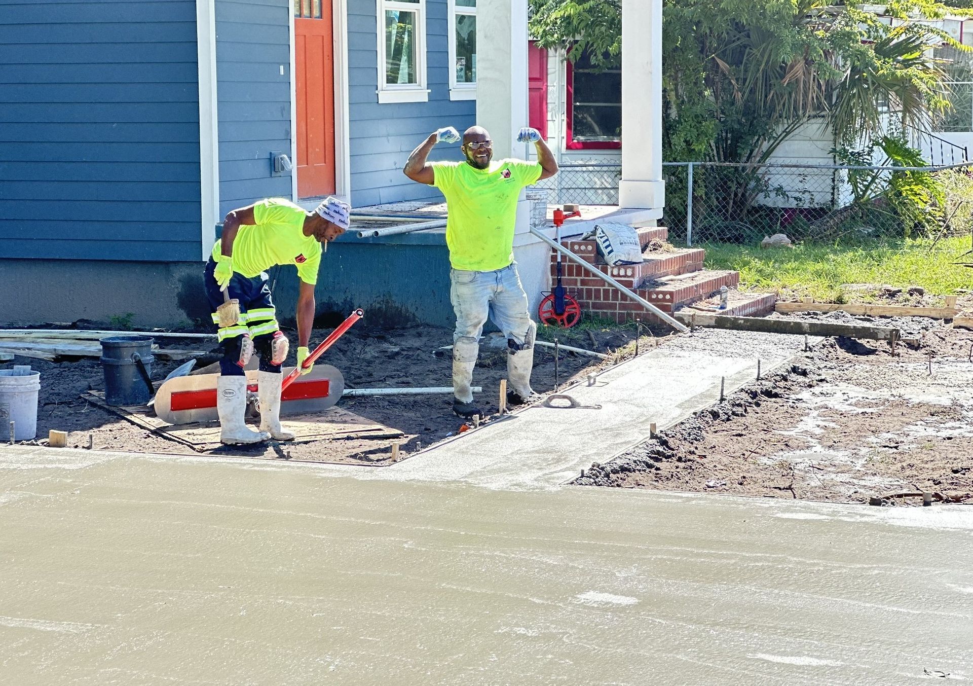 Two workers in safety vests finishing a concrete sidewalk in front of a blue house. One raises arms, smiling.