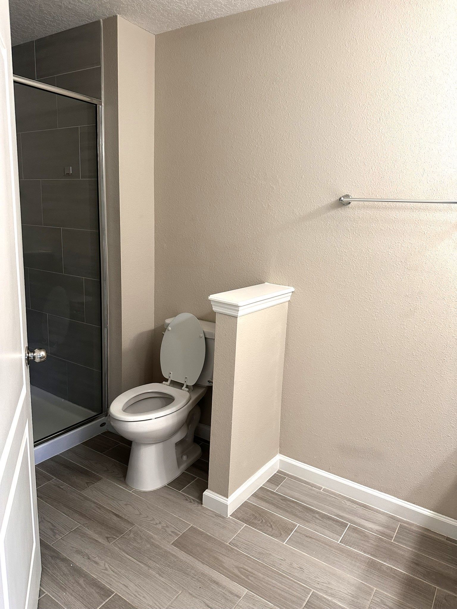 A small bathroom with a toilet, shower, and towel rack. The walls are textured tan, and the floor is gray wood-look tile.
