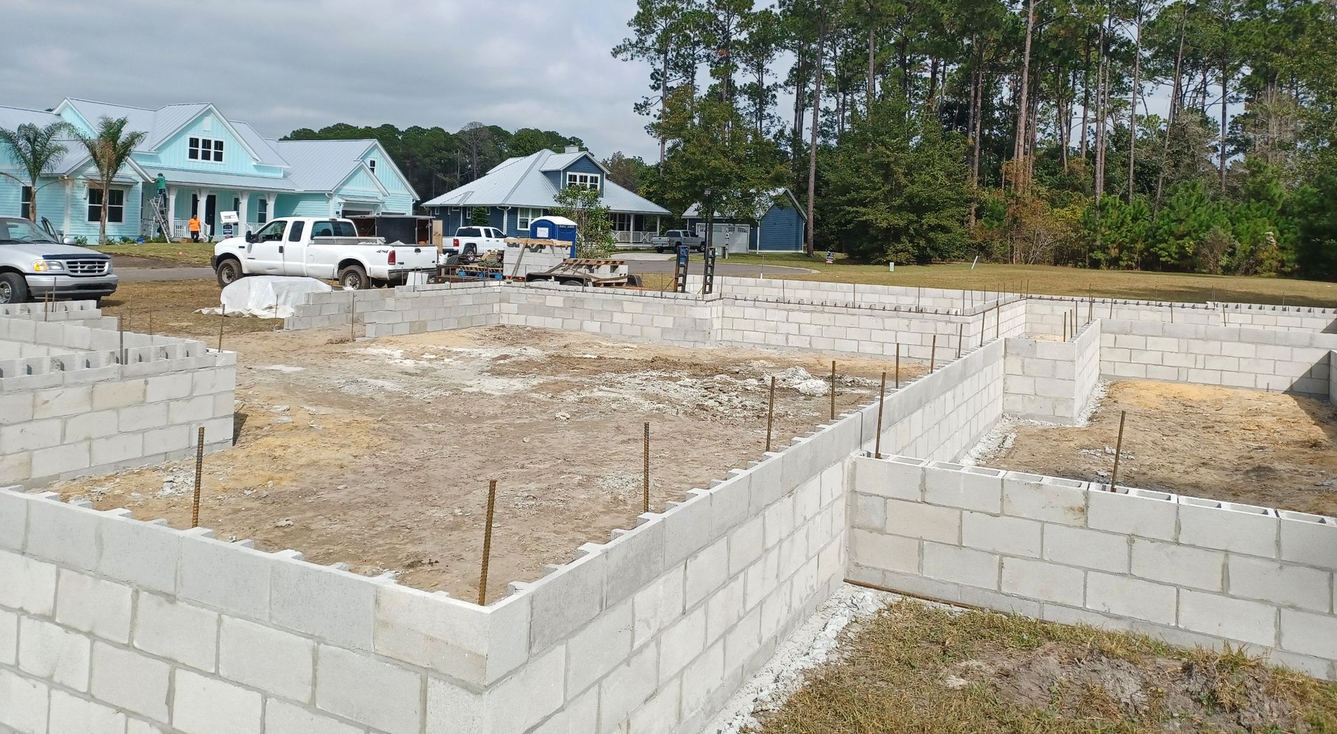 Construction site with gray cinder block foundations. White trucks, houses with blue siding, and trees are in the background.