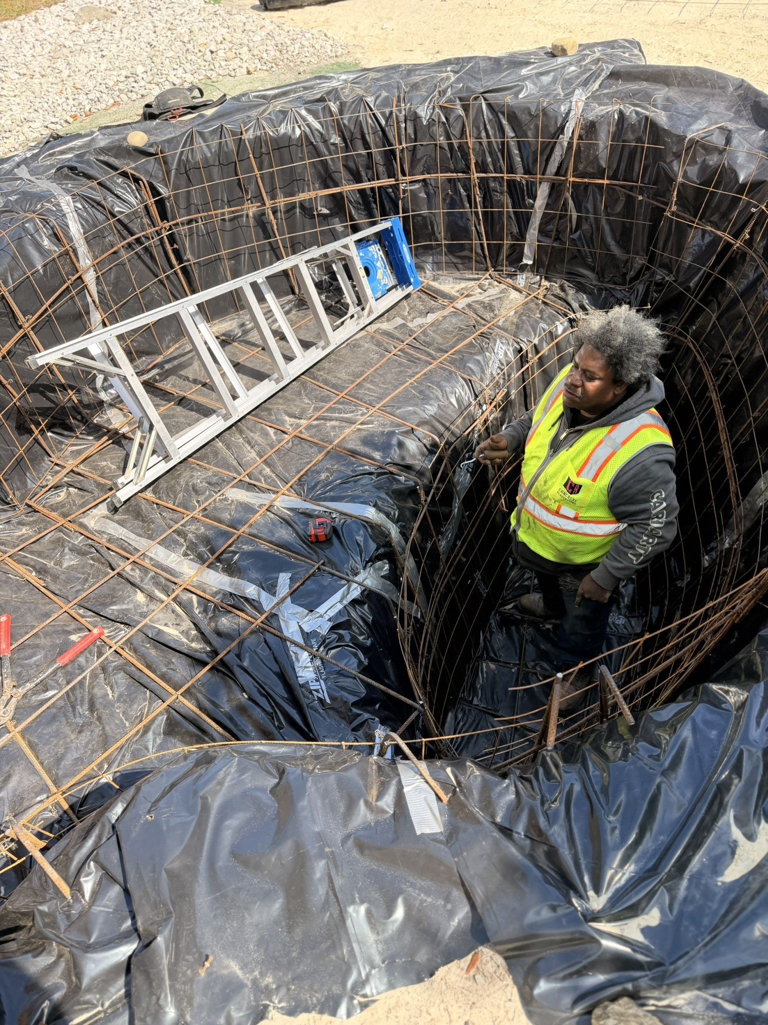 A worker in a high-visibility vest stands inside a circular construction pit lined with black plastic and steel rebar.