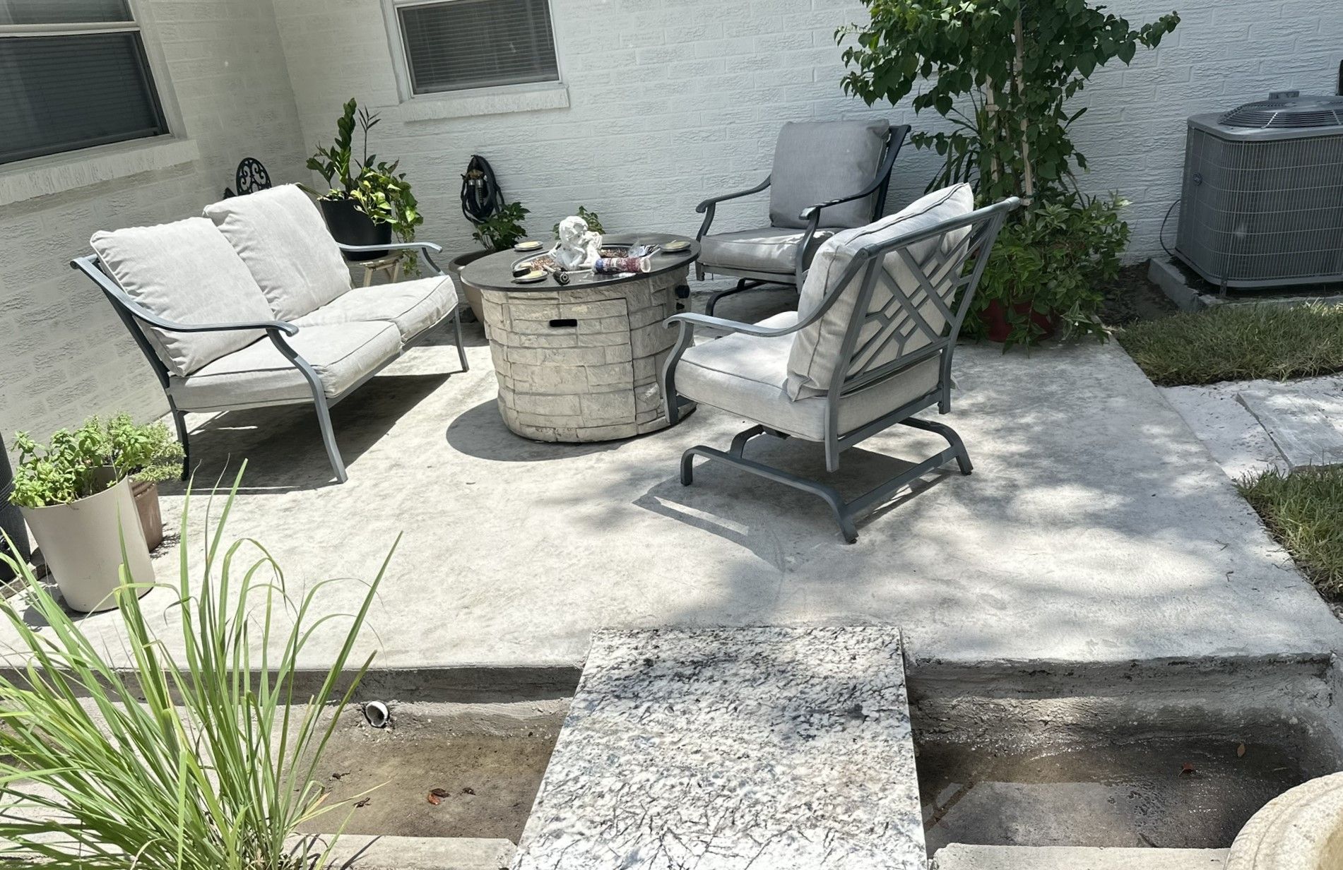 Patio with gray cushioned seating around a stone fire pit.  Small plants and an air conditioning unit are also visible.