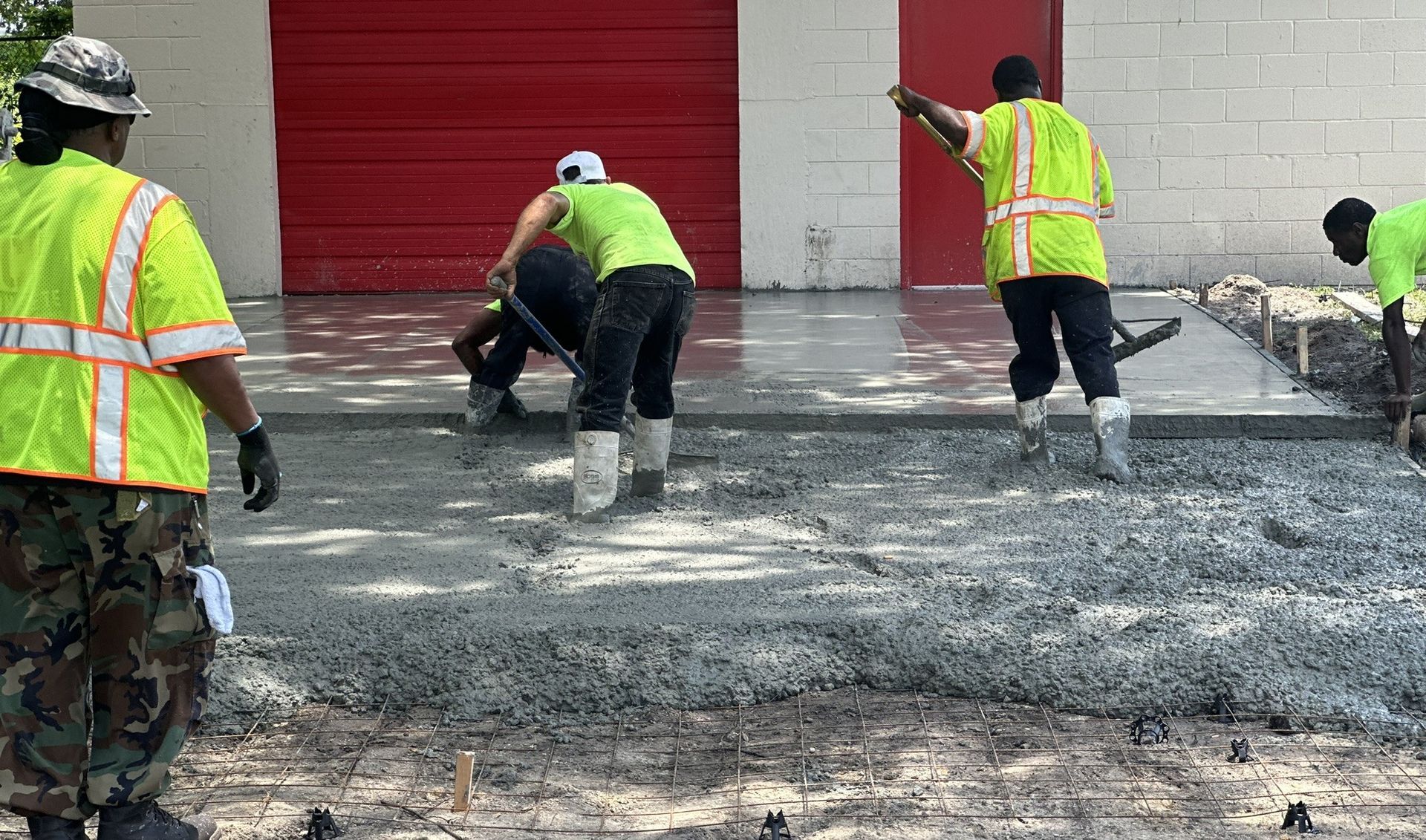 Construction workers pouring concrete on a surface in front of a red door, wearing safety vests and boots.