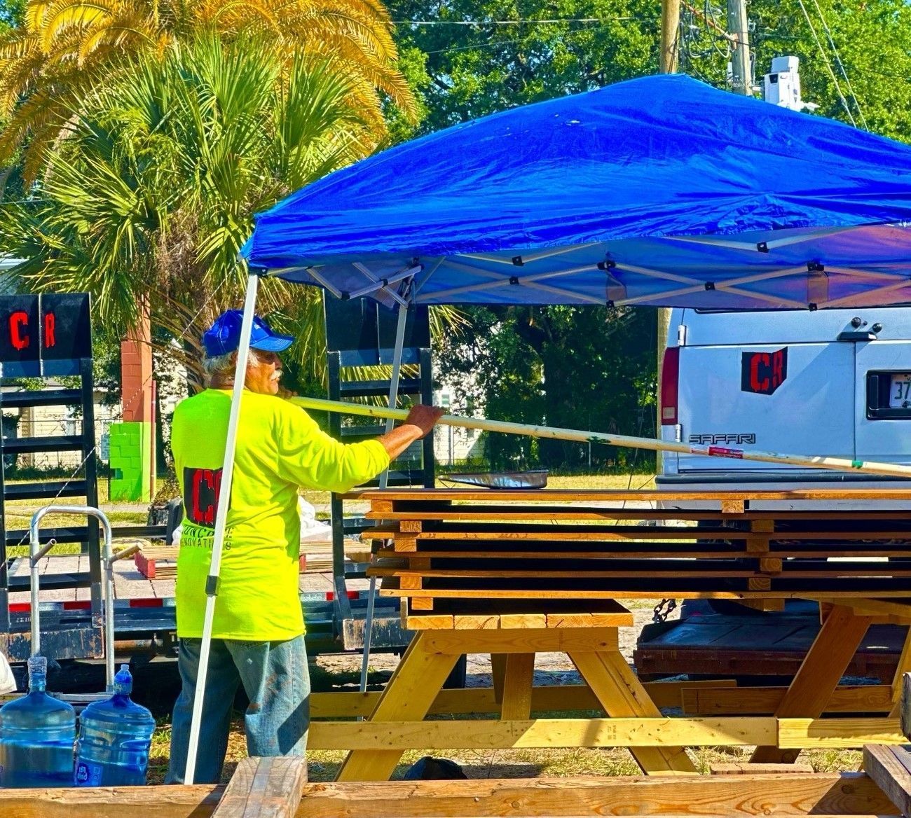 A man in a yellow shirt is standing under a blue tent staining a privacy fence