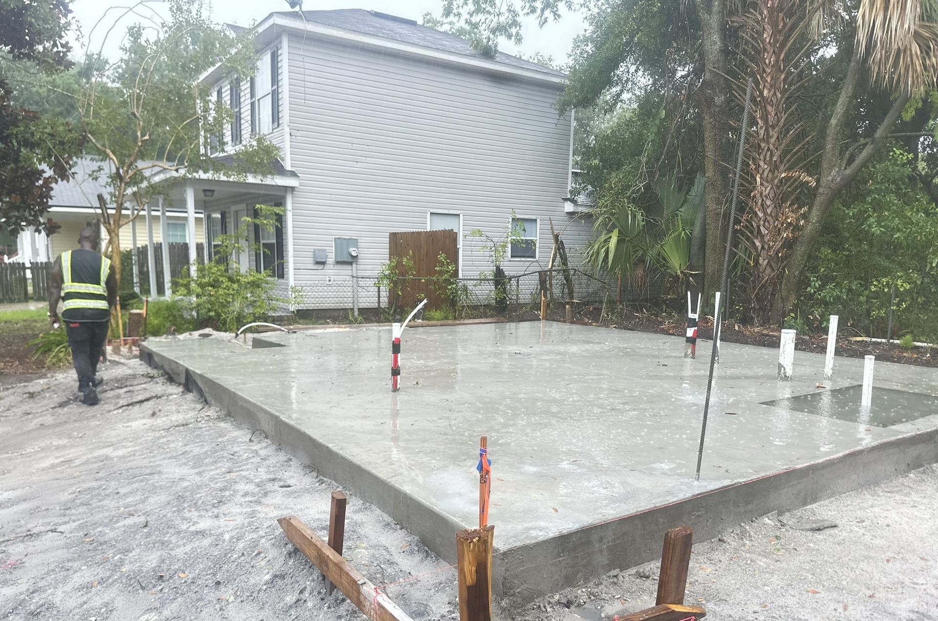 Freshly poured concrete foundation with plumbing pipes, construction worker in vest, residential setting.