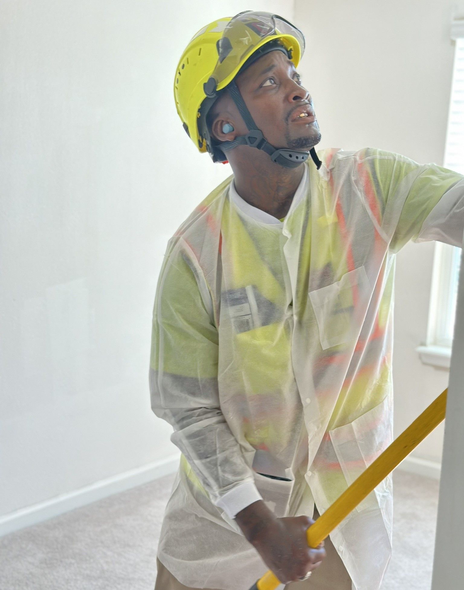 Construction worker in a yellow hard hat and safety vest inspecting a wall, indoors. He is holding a long yellow tool and looking up.
