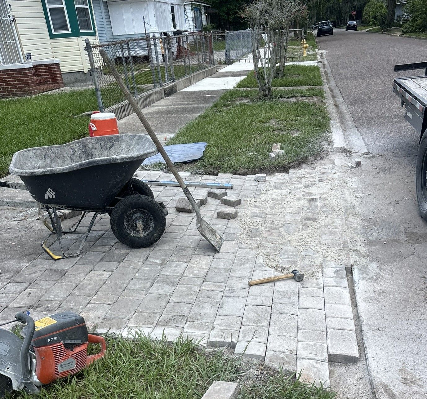 Construction of a brick paver driveway apron. A wheelbarrow, tools, and saw sit beside the work in progress next to a residential street.