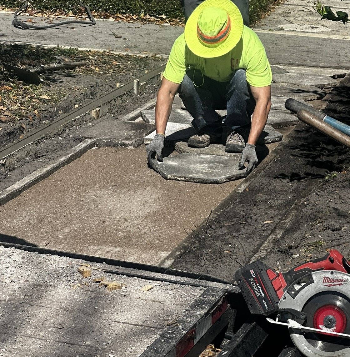A man wearing a yellow hat is kneeling in the dirt installing a paver
