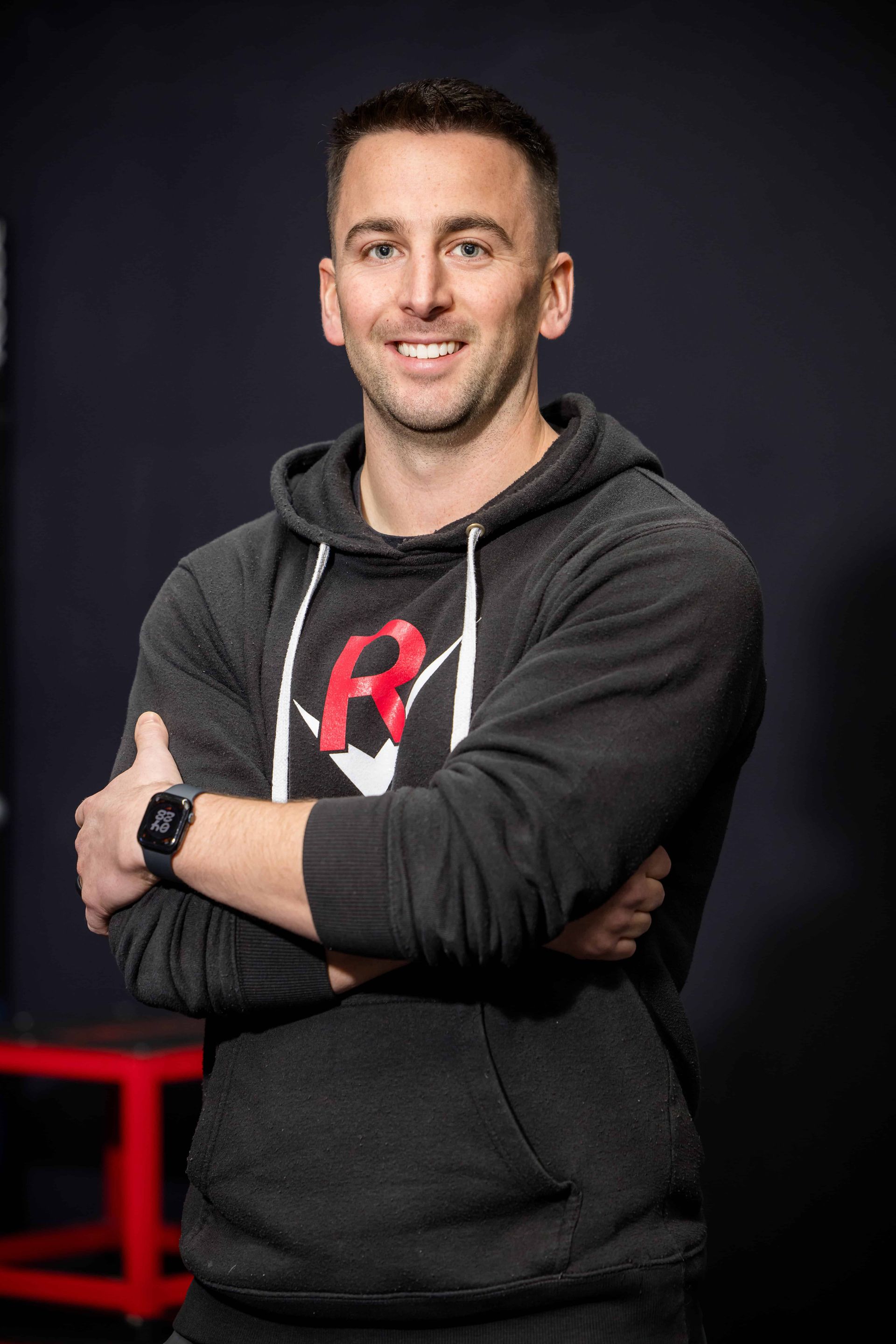 A man wearing a black t-shirt with the letter r on it is standing in a gym with his arms crossed.
