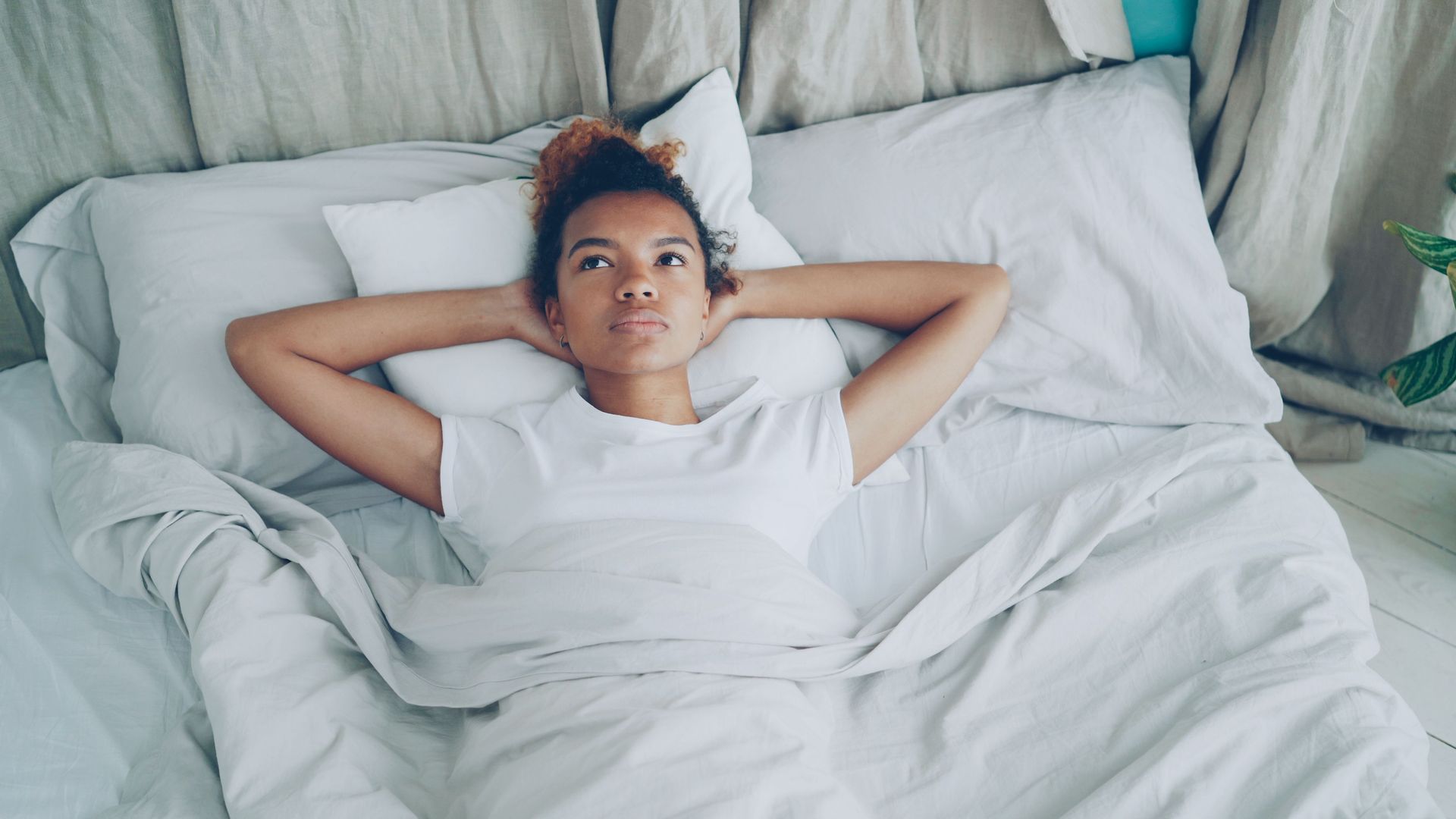 Person lying on a white bed, hands behind head, staring upward in a bright bedroom.