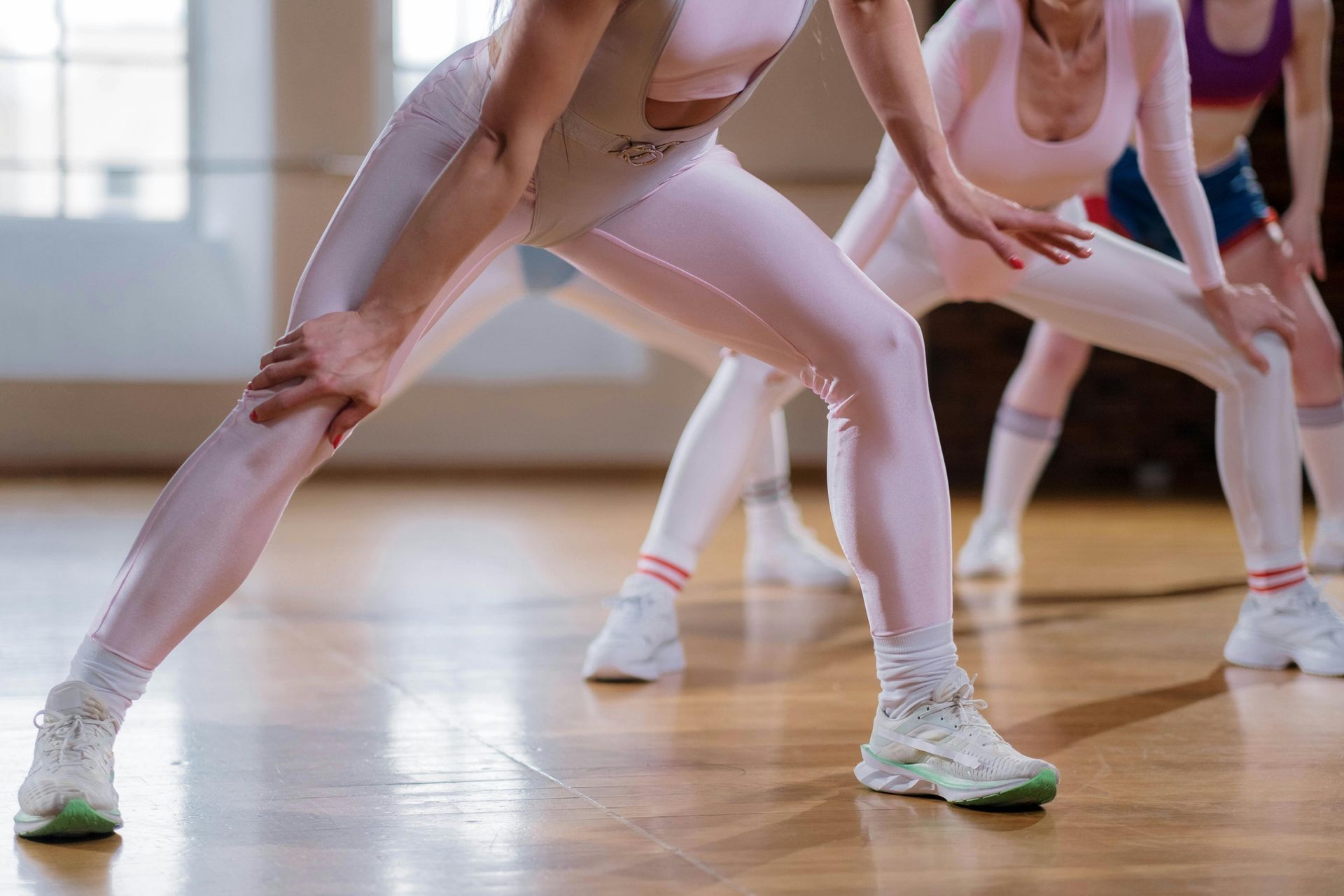 Women in pink workout gear stretching on a wooden floor in a studio.