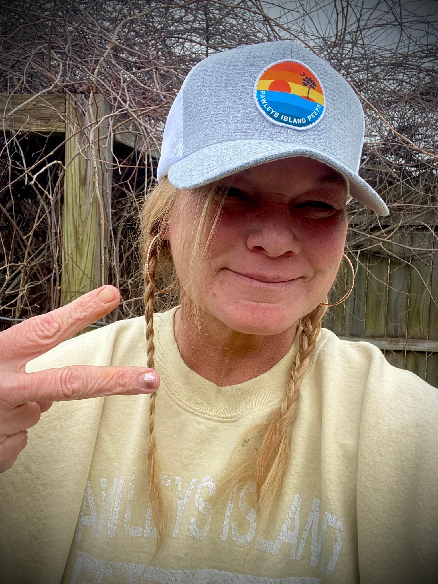 A woman wearing a hat and a yellow shirt is giving a peace sign.