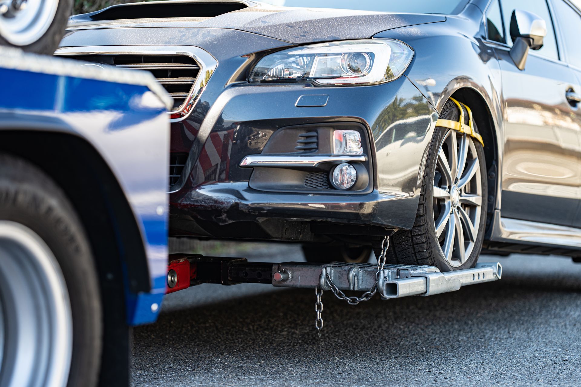 A close-up of a dark sedan being towed by a truck, with the car's front tire secured by a yellow strap and metal lift.