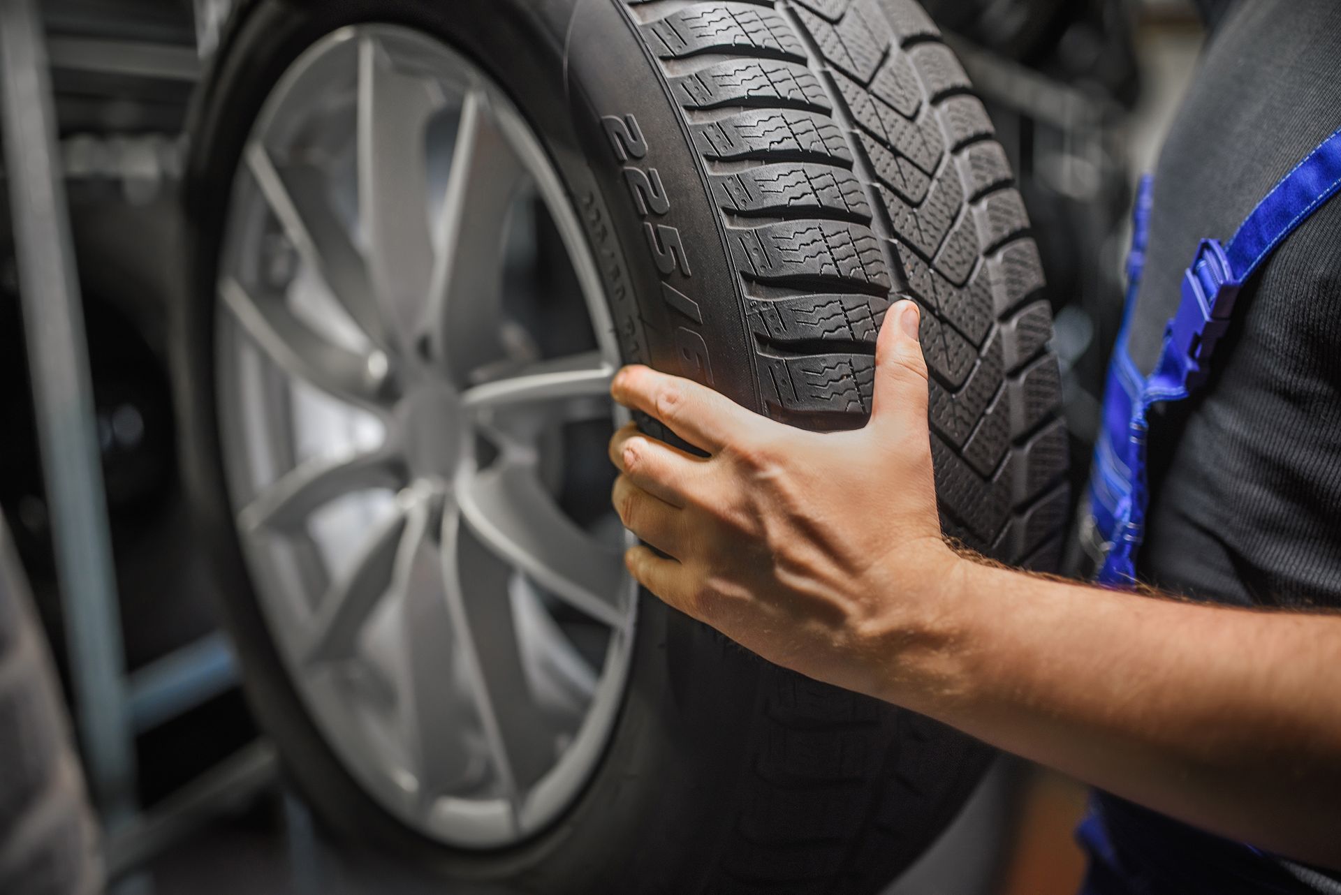 A mechanic in blue overalls holds a car tire with a winter tread pattern, preparing it for mounting on a metal wheel rim.