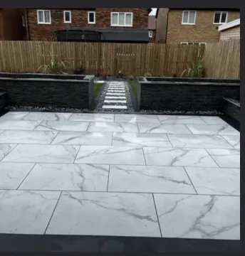 A backyard patio featuring large, white marble-patterned tiles, a stone walkway, and black raised garden beds.