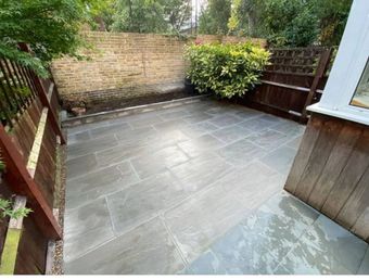 A newly laid grey stone patio in a residential garden, bordered by wooden fencing and a brick wall.