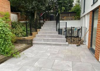 A set of light grey stone stairs leading up through a patio area with black railings and a brick wall on the right.