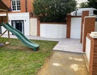 A backyard features a green slide on the grass, a paved patio, and white walls framed by red brick pillars.