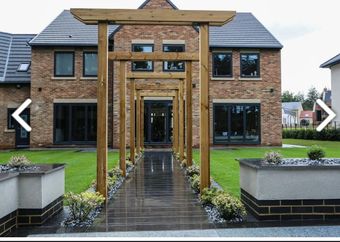 A paved walkway leads through a wooden pergola toward the entrance of a two-story brick house with a green lawn.
