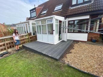 A person stands on a grass lawn next to a grey deck attached to a white conservatory on the back of a brick house.