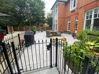 A paved patio area behind a red brick building, featuring outdoor furniture, a grill, and a black metal gate in front.