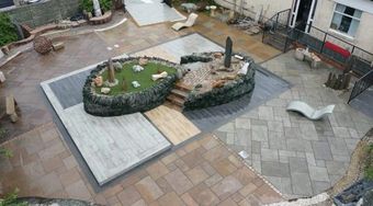 An elevated view of a patio garden featuring mixed paving, a central rockery with a grass section, and lounge seating.