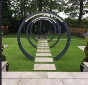 A garden path with three sequential, circular metal archways leading to a tree-lined background on a manicured lawn.