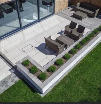 High-angle view of a modern backyard patio with light stone tiles, outdoor seating, and an L-shaped raised garden planter.