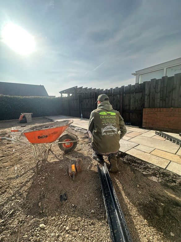 A beige stone patio extends from a modern brick house, with a wet surface reflecting the sky, beside a grassy lawn.
