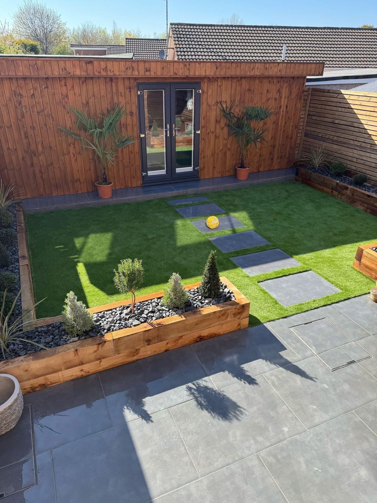 Backyard patio with artificial lawn, stone pavers, wooden shed, and potted plants in bright sunlight