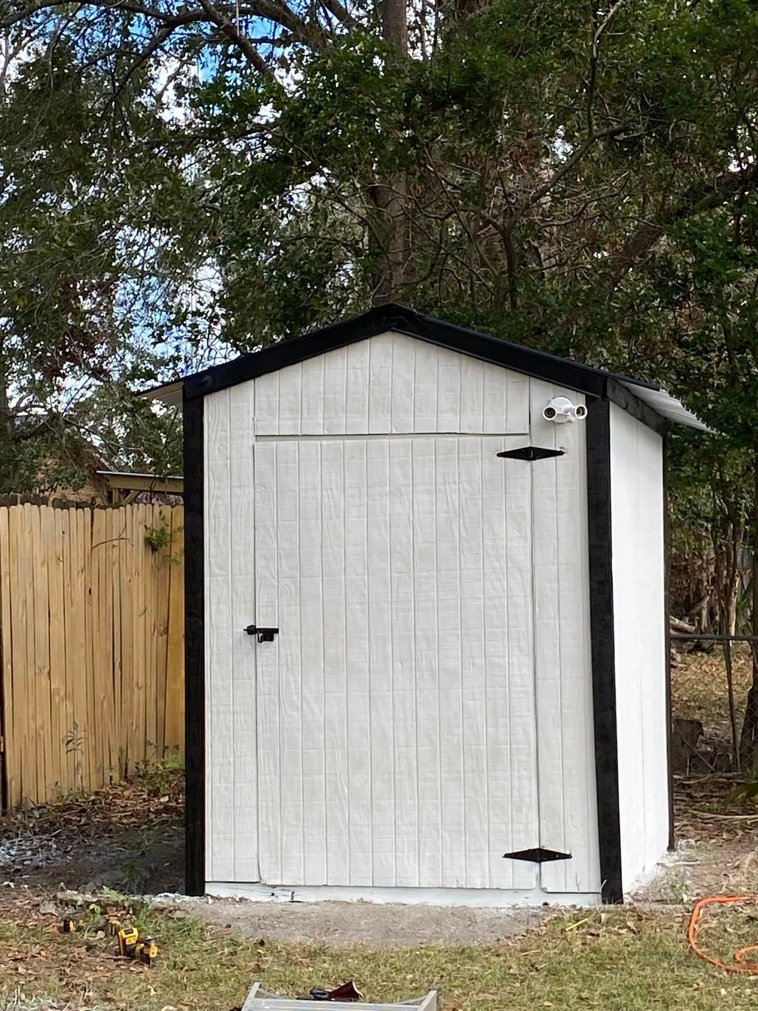 A white shed with a black trim is sitting in the grass next to a wooden fence.