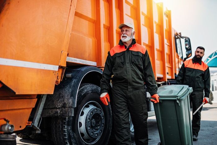 Two sanitation workers near orange garbage truck, one holding green trash can.