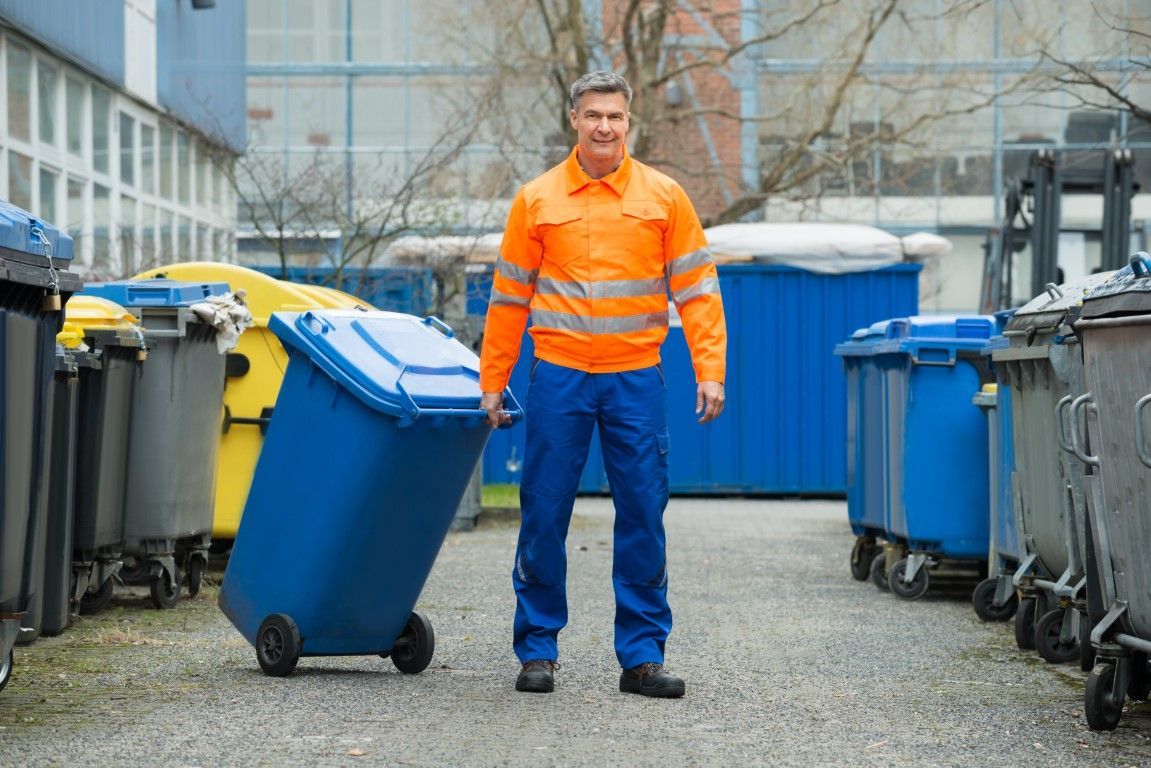 Man in orange safety vest and blue pants pulls a blue trash bin between rows of bins.