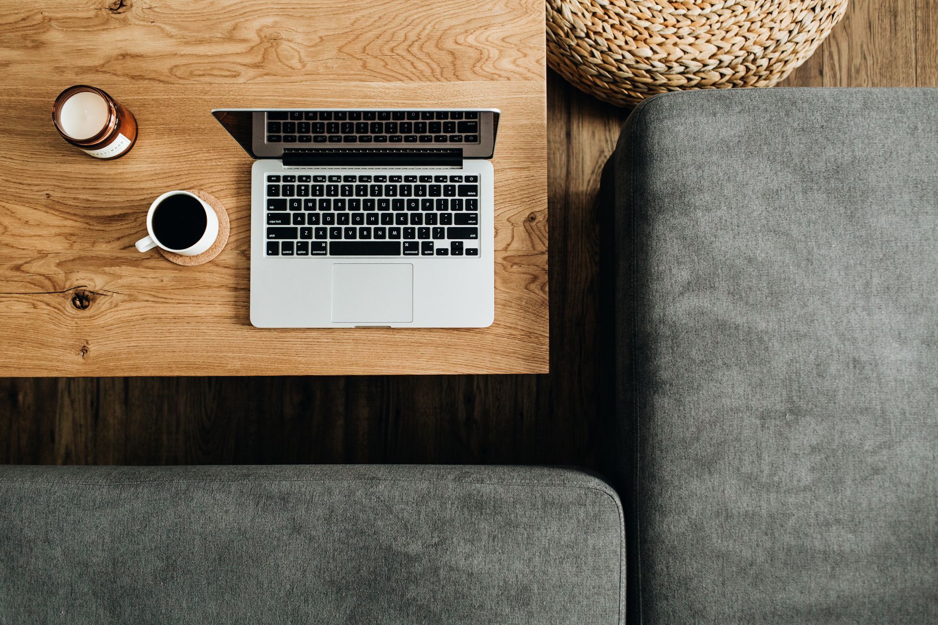 Laptop open on a wooden coffee table with a coffee cup and candle; next to a gray sofa and wicker basket.